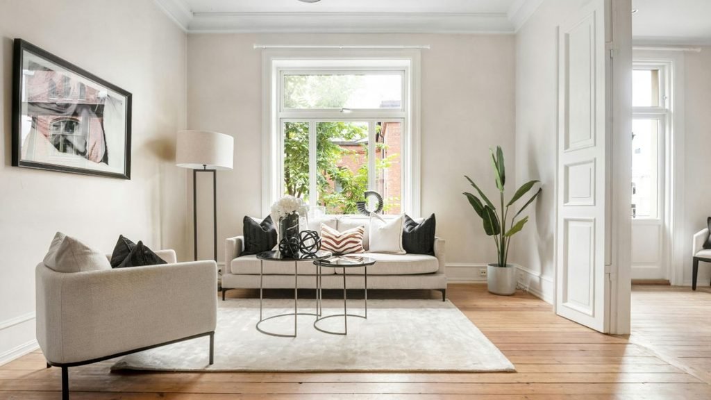 text: Bright apartment living room with a cream sofa, nesting coffee tables, a tall plant and wooden floors.