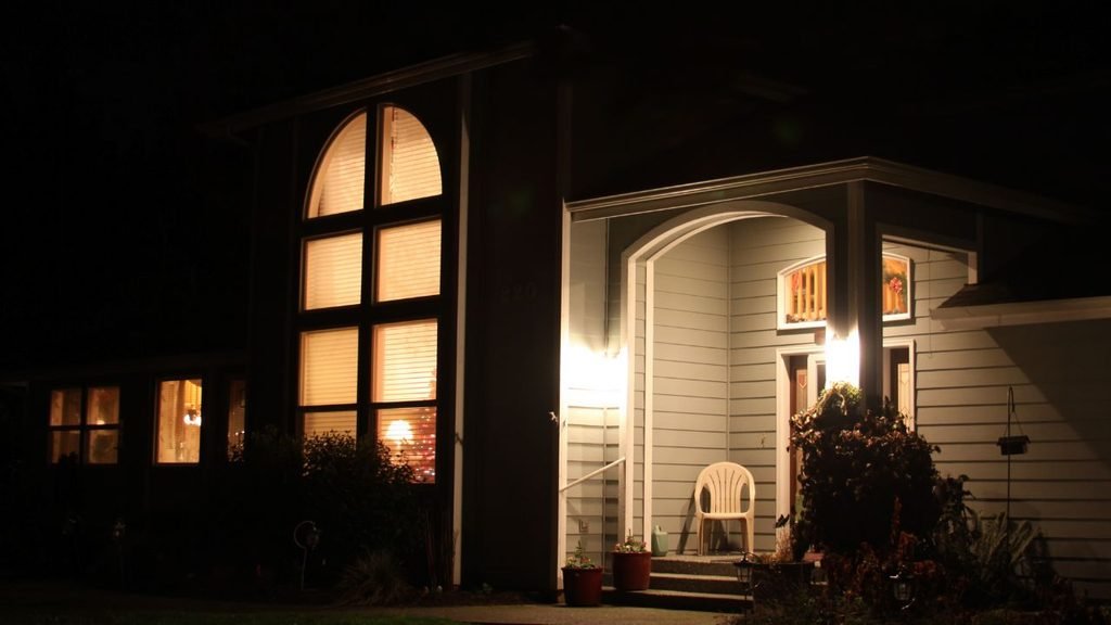 Arched front porch glowing at night with a wall light, a white plastic chair and warmly lit windows behind it