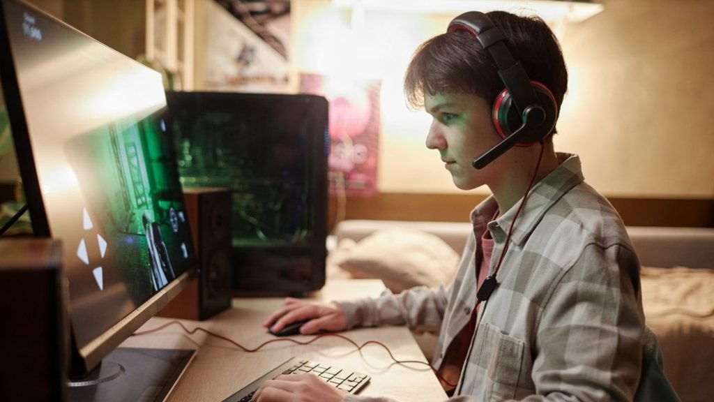 Teen with a headset playing at a compact bedroom desk with a monitor and speakers, a simple gaming setup in a room