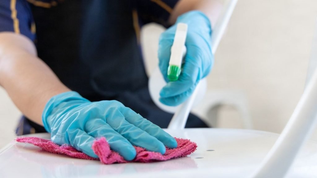 Hands in teal gloves polishing a chrome bathroom faucet with a yellow cloth and spray cleaner