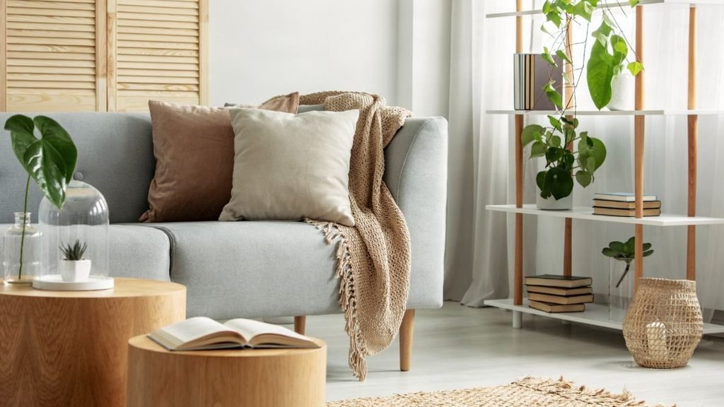 Gray sofa with a knit blanket and soft pillows beside round oak side tables and a shelf of plants.