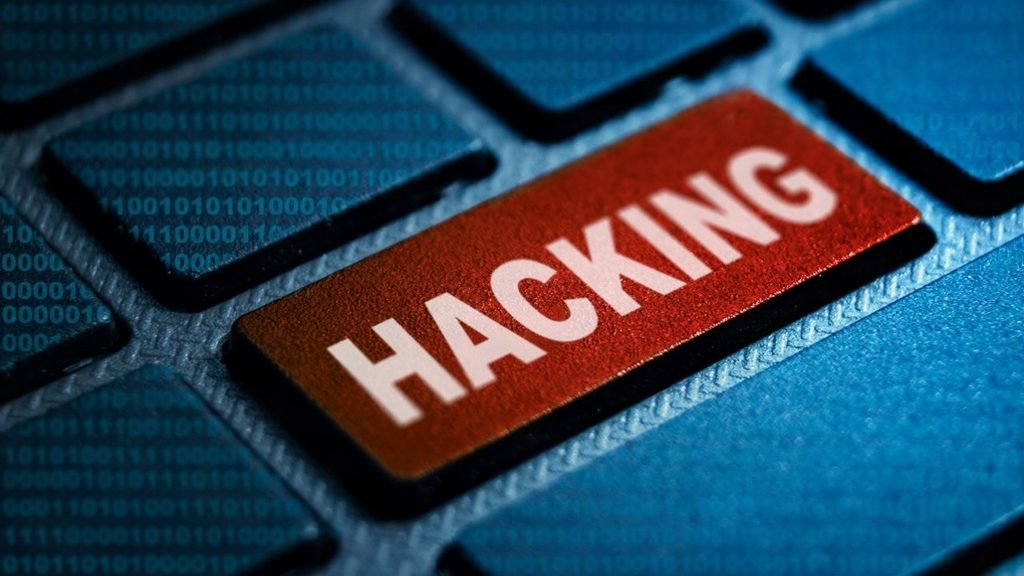 Red keyboard key labeled “HACKING” on a blue keyboard with binary code in the background