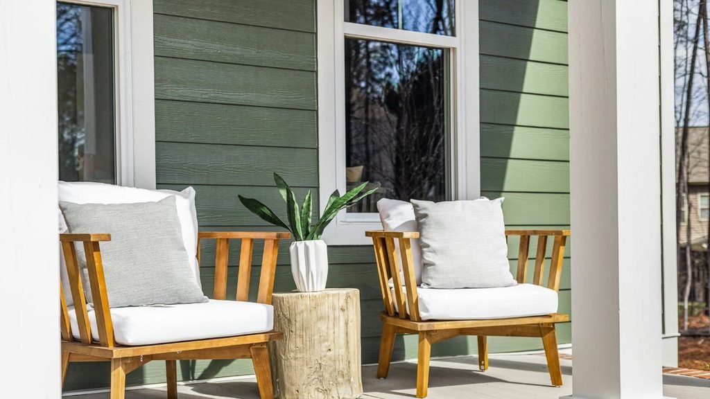 Two wood-frame porch chairs with white cushions and gray pillows around a log side table with a snake plant, against green siding