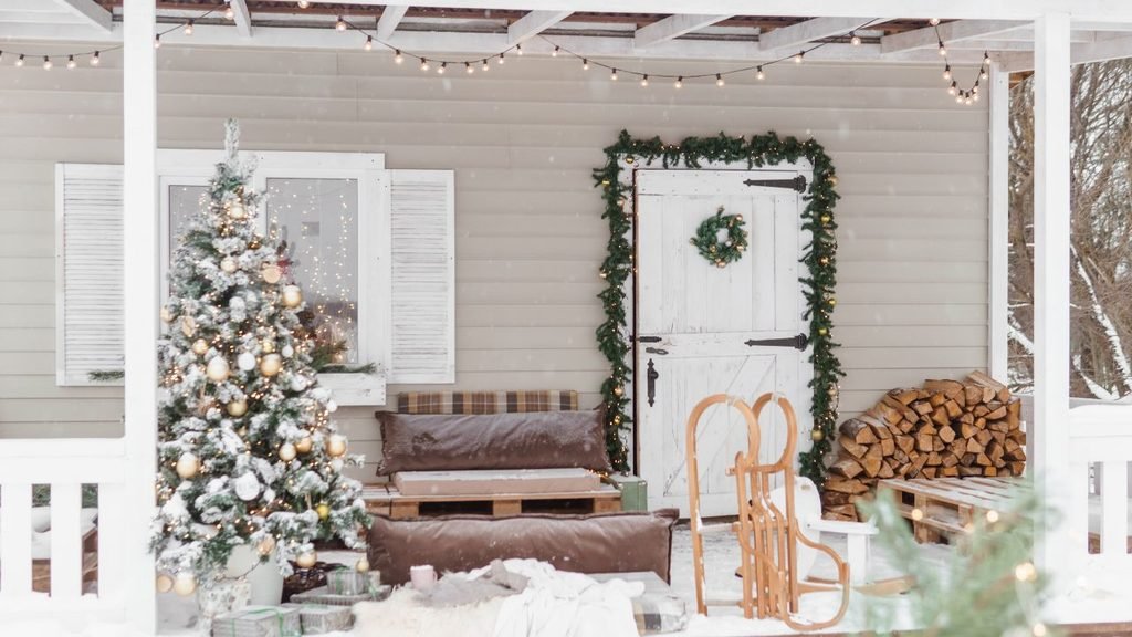 Cozy snowy porch idea for houses with a flocked Christmas tree, white barn-style door framed by garland, string lights and stacked firewood