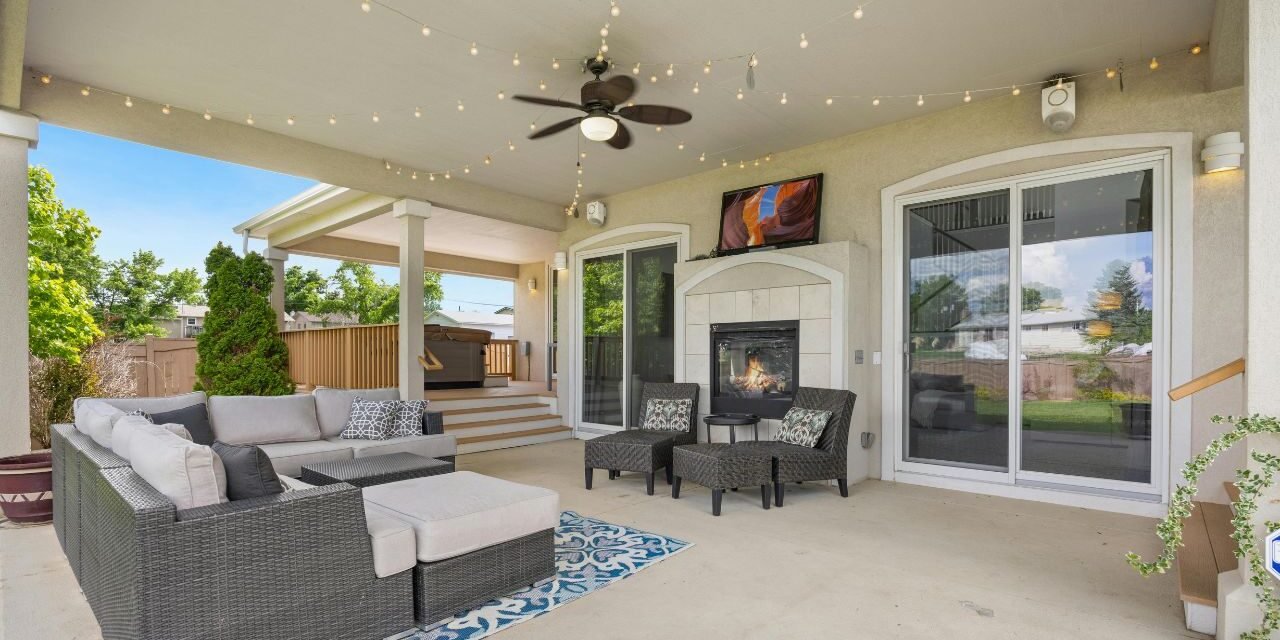 3: Spacious covered patio with a wicker sectional, gas fireplace, ceiling fan and string lights, with steps leading down to a backyard deck