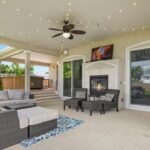 3: Spacious covered patio with a wicker sectional, gas fireplace, ceiling fan and string lights, with steps leading down to a backyard deck