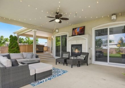 3: Spacious covered patio with a wicker sectional, gas fireplace, ceiling fan and string lights, with steps leading down to a backyard deck