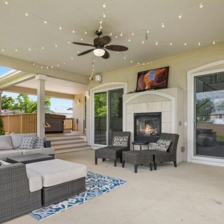 3: Spacious covered patio with a wicker sectional, gas fireplace, ceiling fan and string lights, with steps leading down to a backyard deck