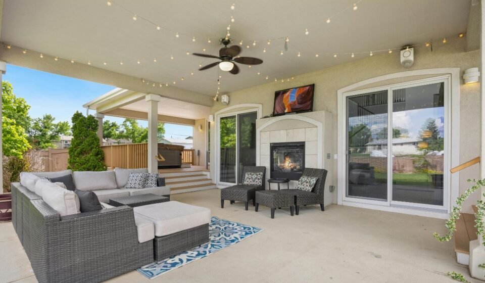 3: Spacious covered patio with a wicker sectional, gas fireplace, ceiling fan and string lights, with steps leading down to a backyard deck