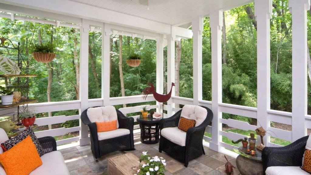 Screened porch with black wicker chairs, orange accent pillows and a stone floor facing tall trees, a good fit for porch ideas for houses with a view