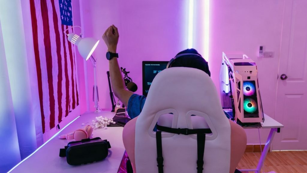 Gamer celebrating in a white gaming chair in a room with purple LED strip lighting, a white PC case and a VR headset on the desk