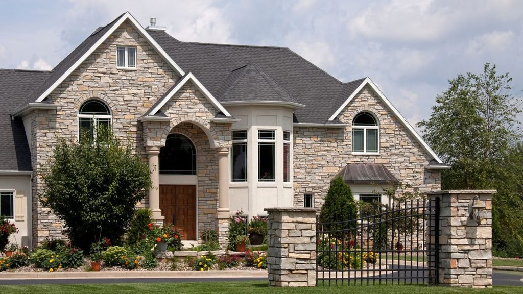 Stone house architecture with an arched wooden front door, bay window and wrought iron gate framed by flower beds