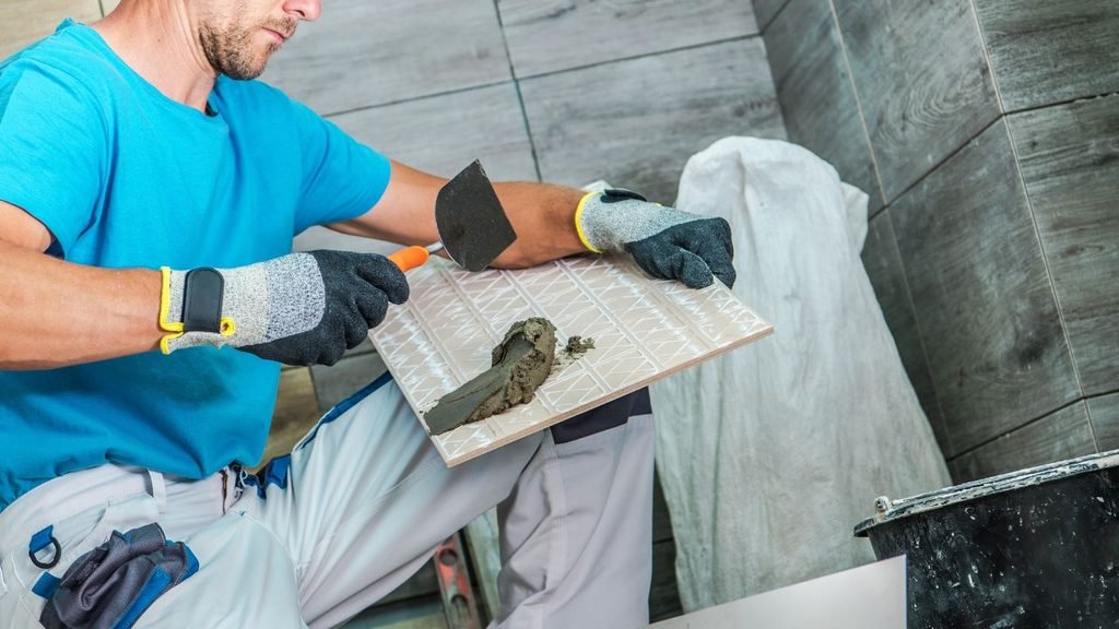 Close-up of gloved hands applying adhesive to a ceramic tile with a trowel before bathroom wall installation