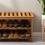 Wooden slatted shoe rack organizer bench with two shelves holding men's boots and leather shoes beside a potted snake plant